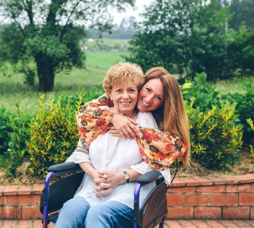 Alltagsheldin umarmt eine lächelnde Seniorin im Rollstuhl im Garten – Seniorenbetreuung von Alltagshelden 24